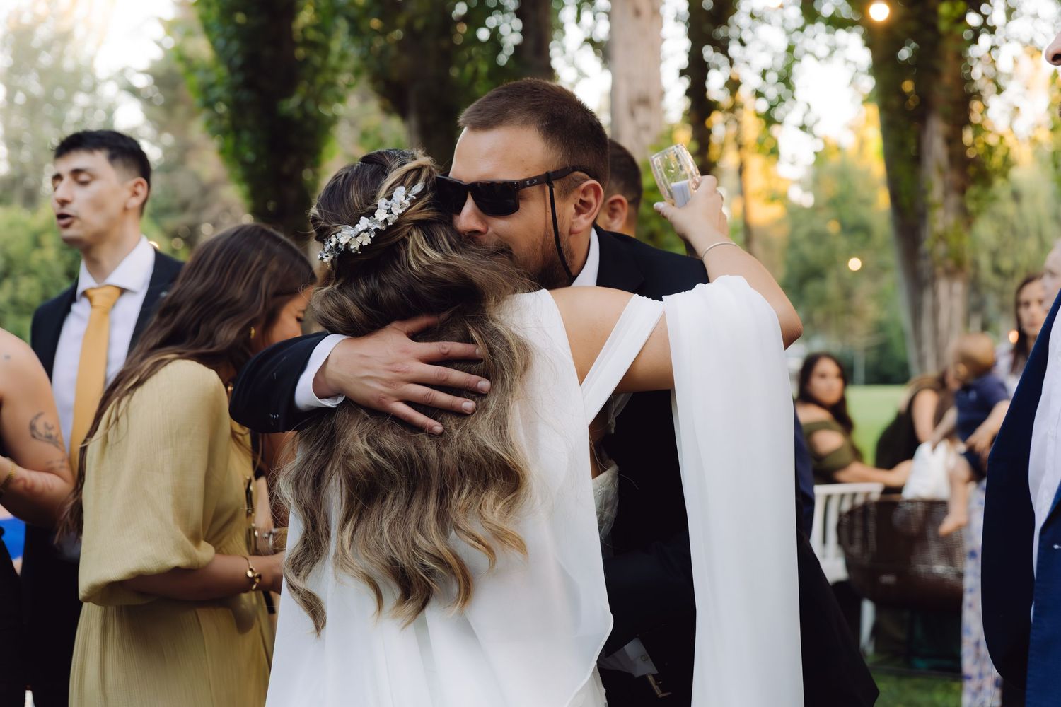 Bride wearing floral crown receives warm embrace from sunglasses-wearing groom at outdoor wedding celebration.