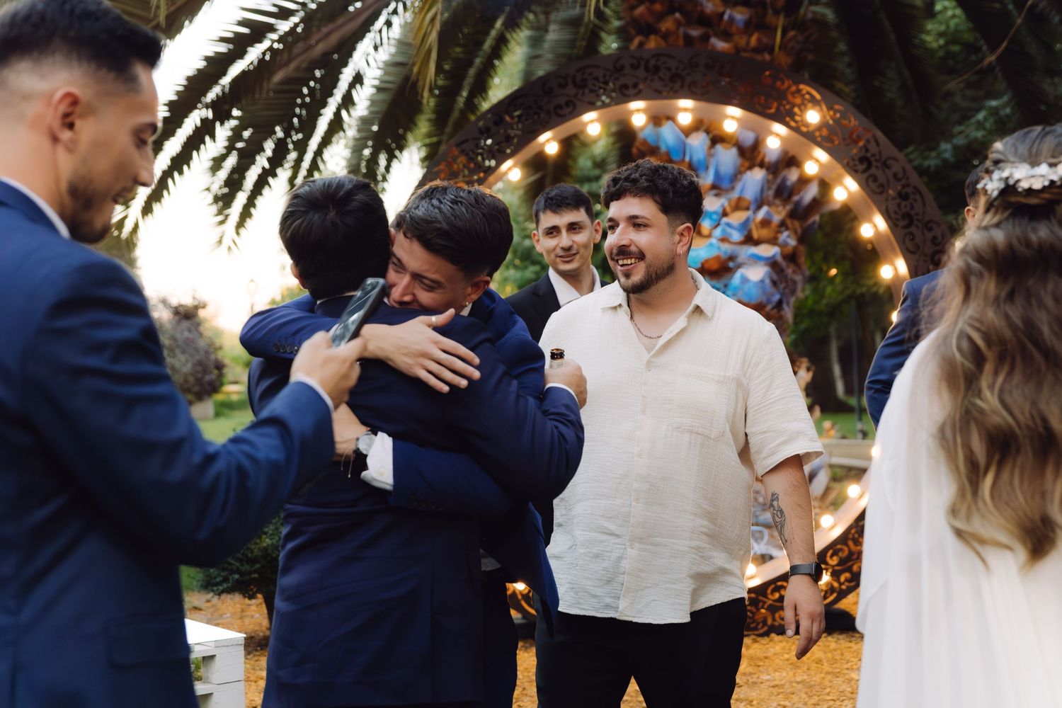 Wedding guests sharing joyful hugs and laughter near decorative arch at outdoor reception venue.