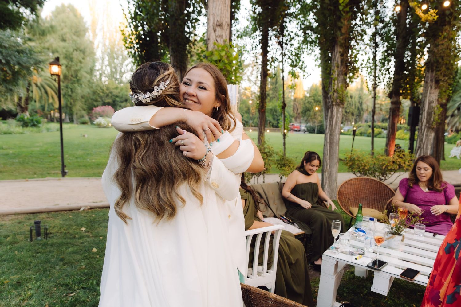 Bride in white gown warmly embracing female wedding guest at elegant outdoor garden reception.