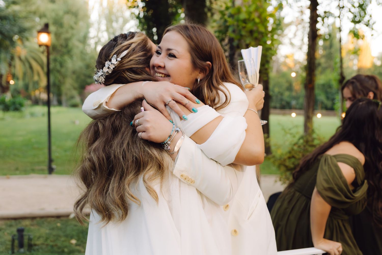 Two women in white dresses sharing a heartfelt hug at an outdoor wedding celebration.