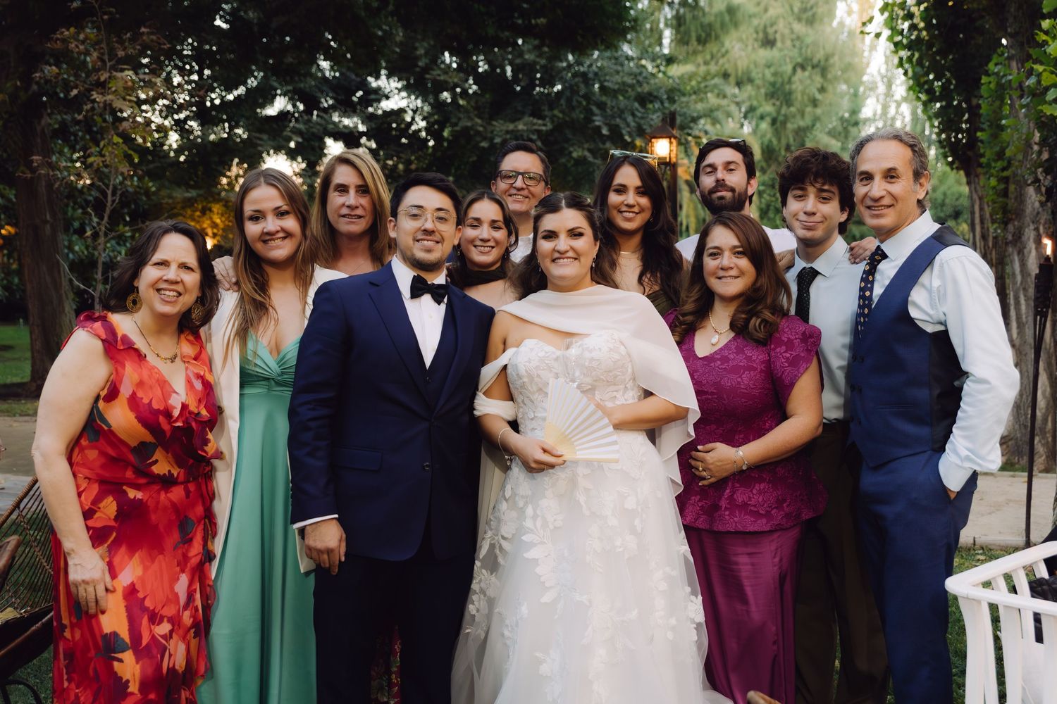 Wedding couple poses with family group photo outdoors surrounded by lush green trees.