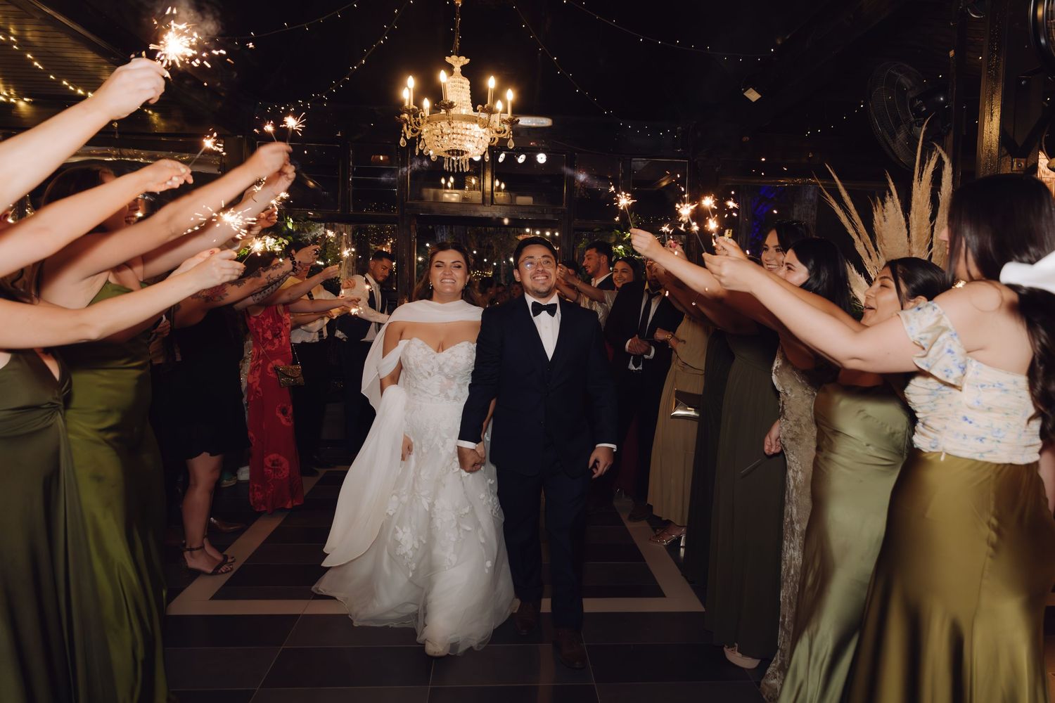 Bride in off-shoulder gown and groom in tuxedo walk through sparkler exit lined with wedding guests.