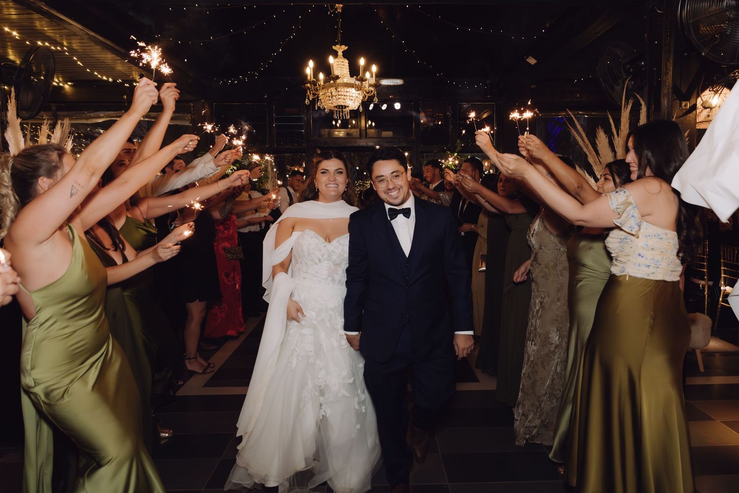 Smiling newlyweds celebrate their sparkler grand exit surrounded by bridesmaids in olive green gowns.