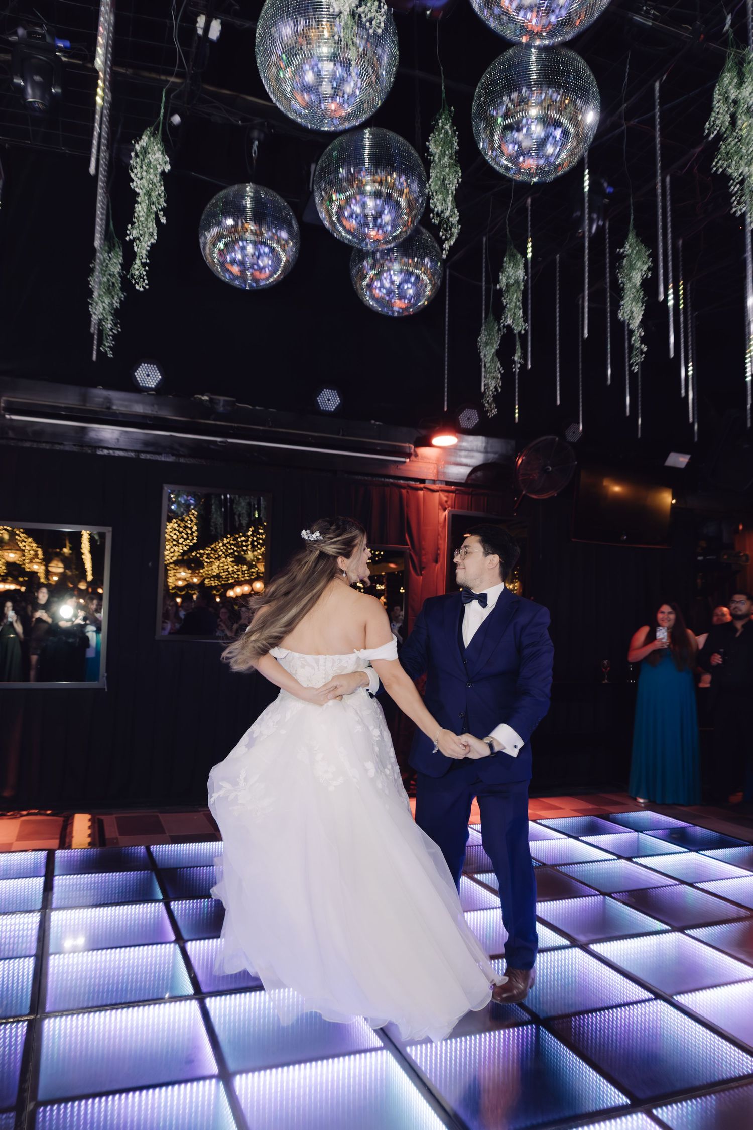 Newlyweds dance joyfully on illuminated LED dance floor beneath hanging disco balls at wedding reception.