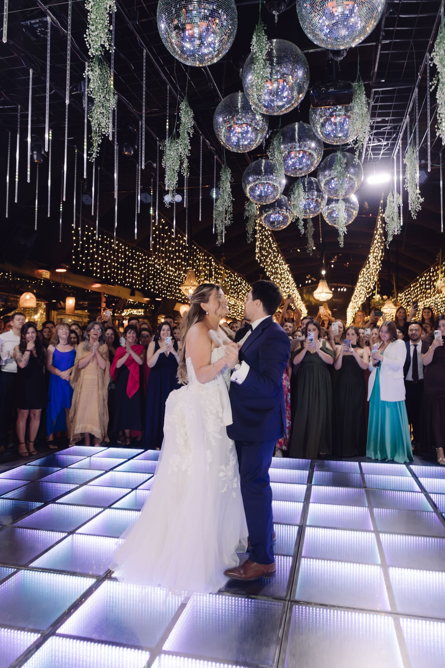 Bride and groom share first dance on glowing dance floor under disco balls while guests applaud.