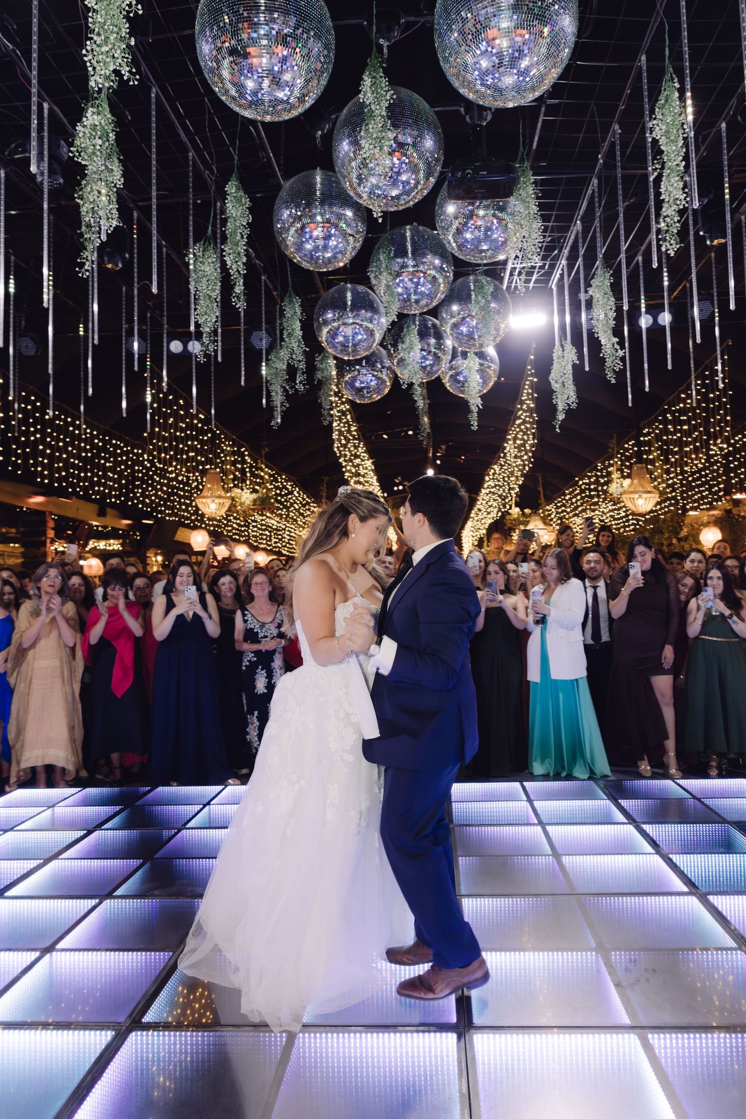 Newlyweds embrace during first dance under mirrored disco balls and twinkling lights at wedding reception.