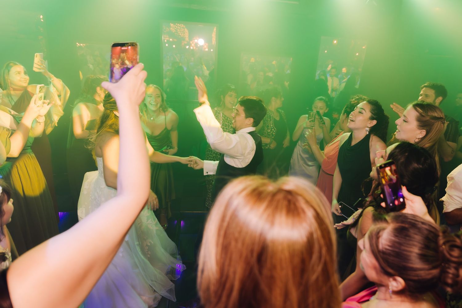 Lively wedding reception dance floor with guests celebrating under green lights while someone records on a smartphone.