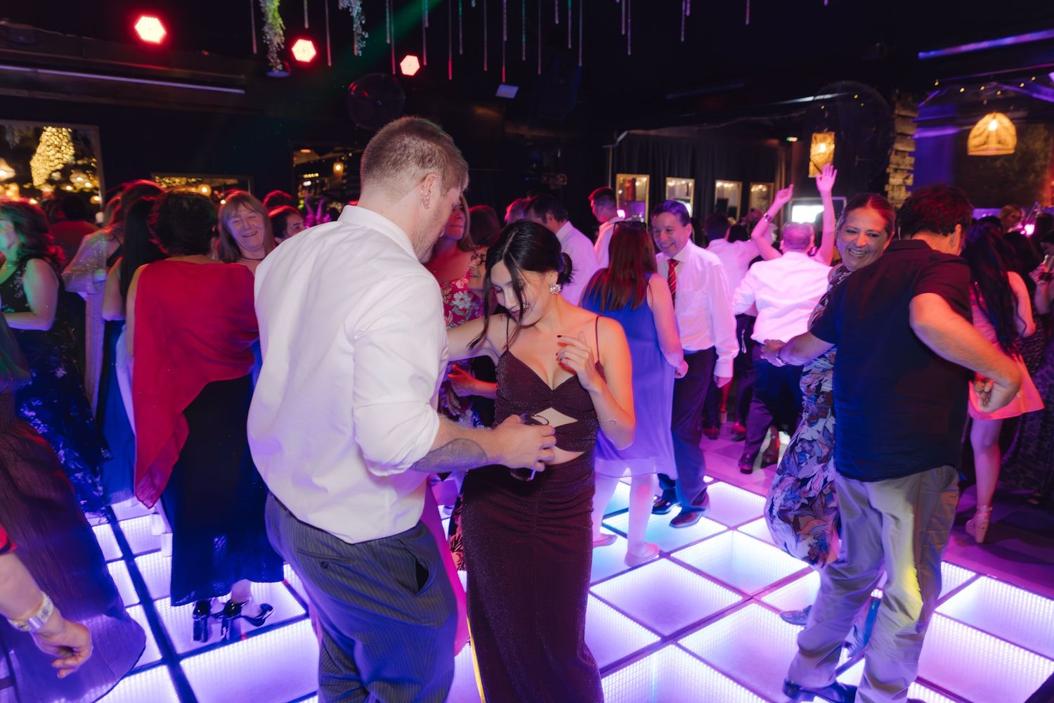 Crowd of elegantly dressed guests dancing on illuminated dance floor at a nightclub party.