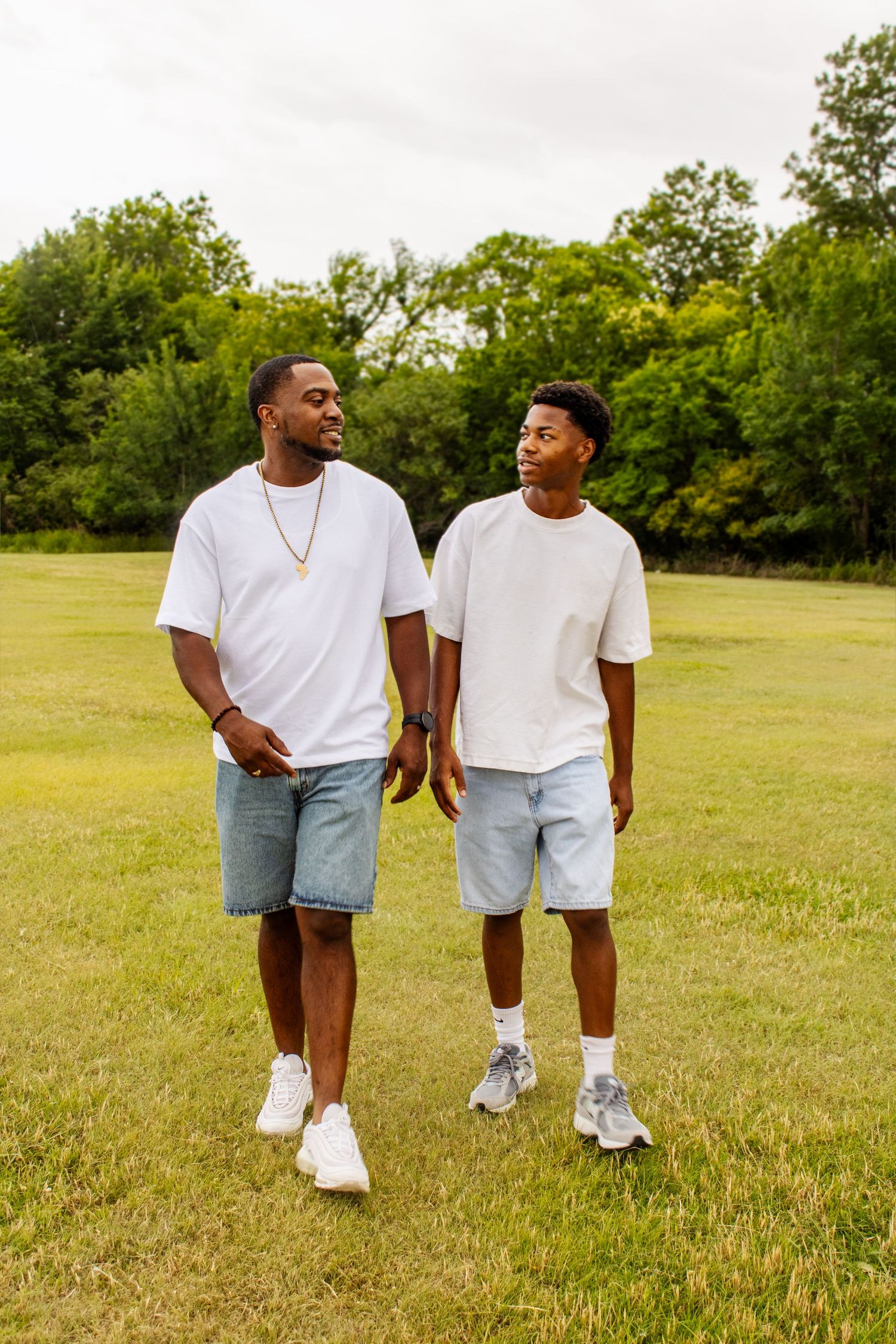 Two Black men in matching white tees and denim shorts walk together on a green grass field.