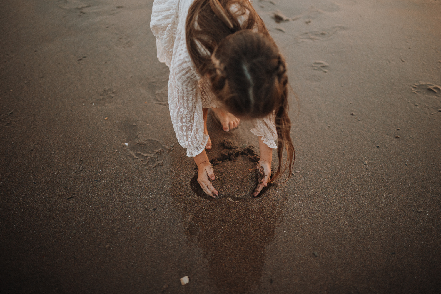 A young girl in a white dress bends down to draw in wet sand on a beach.