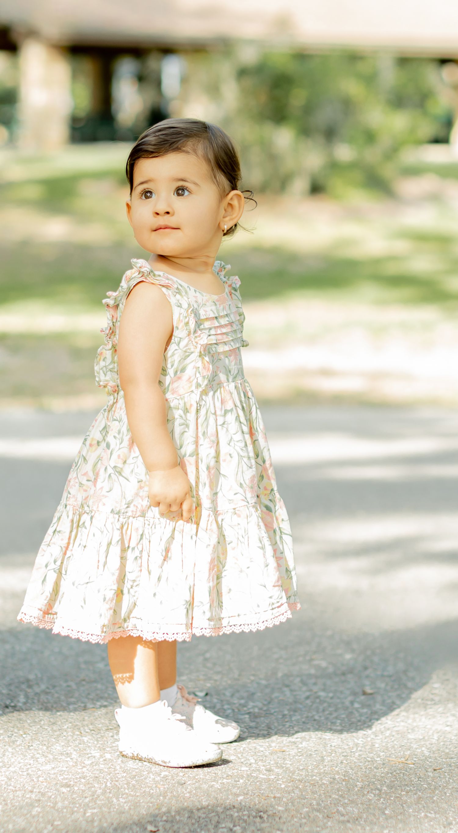 A toddler wearing a white floral sundress plays on a sunlit outdoor path in summer.