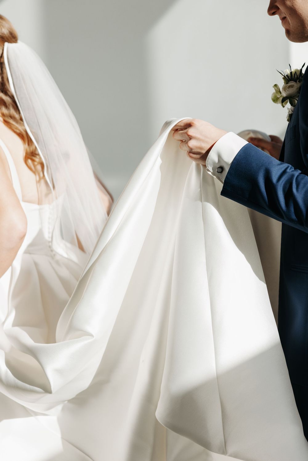 Groom in navy suit adjusts bride's white wedding dress train indoors, showcasing elegant bridal gown details.