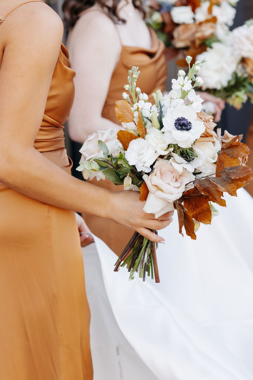 Bridesmaids in golden satin dresses holding elegant fall wedding bouquets with white anemones and roses.
