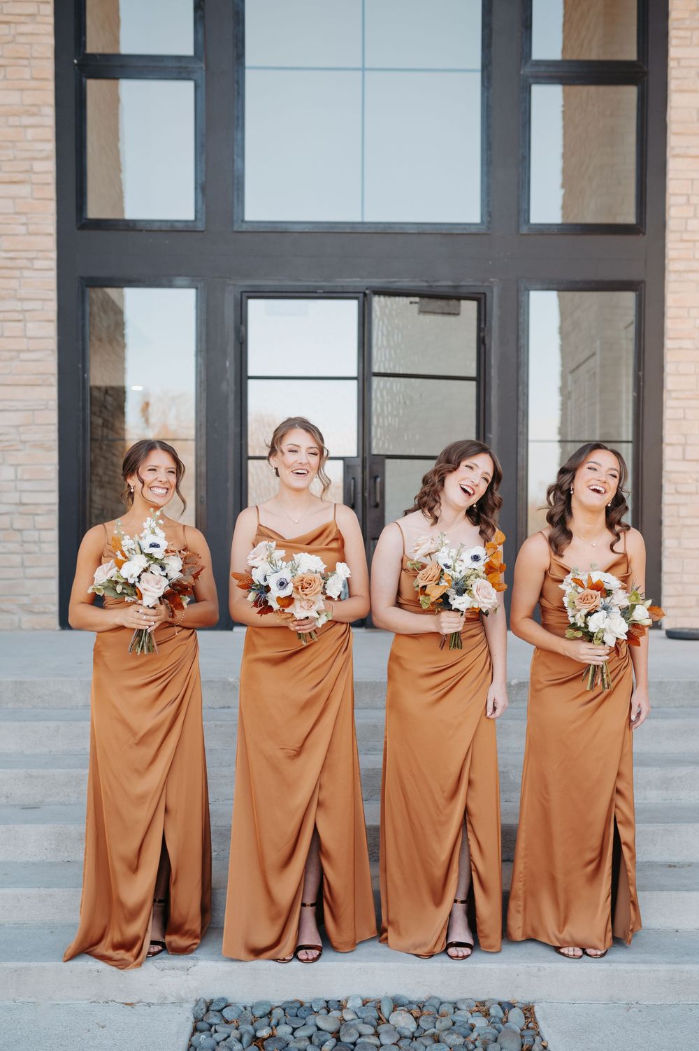 Four bridesmaids in rust-orange satin gowns laughing and holding floral bouquets outside a modern building.