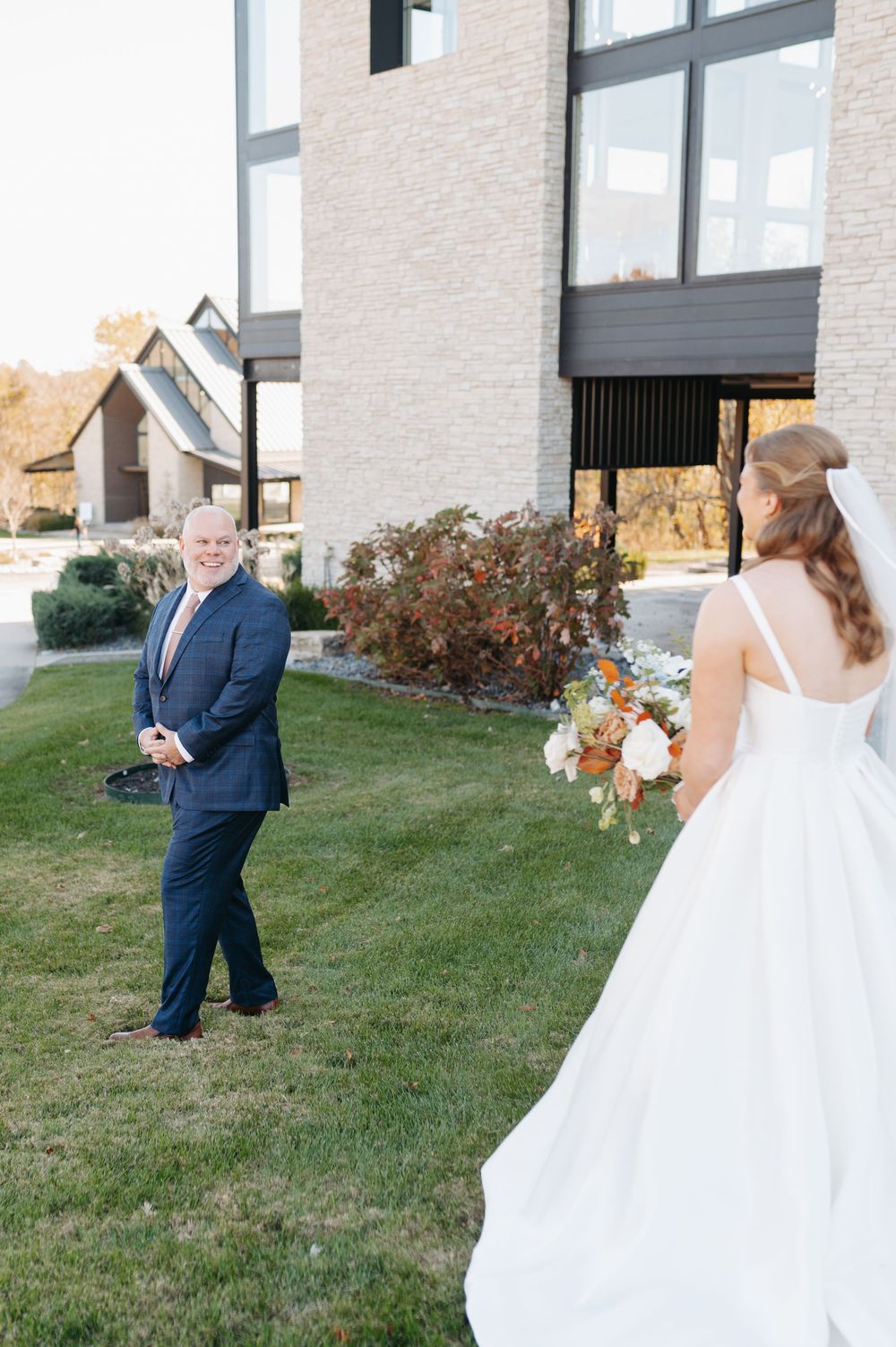 A bride in a white gown holds a colorful bouquet during an emotional first look with a man in a navy suit outdoors.