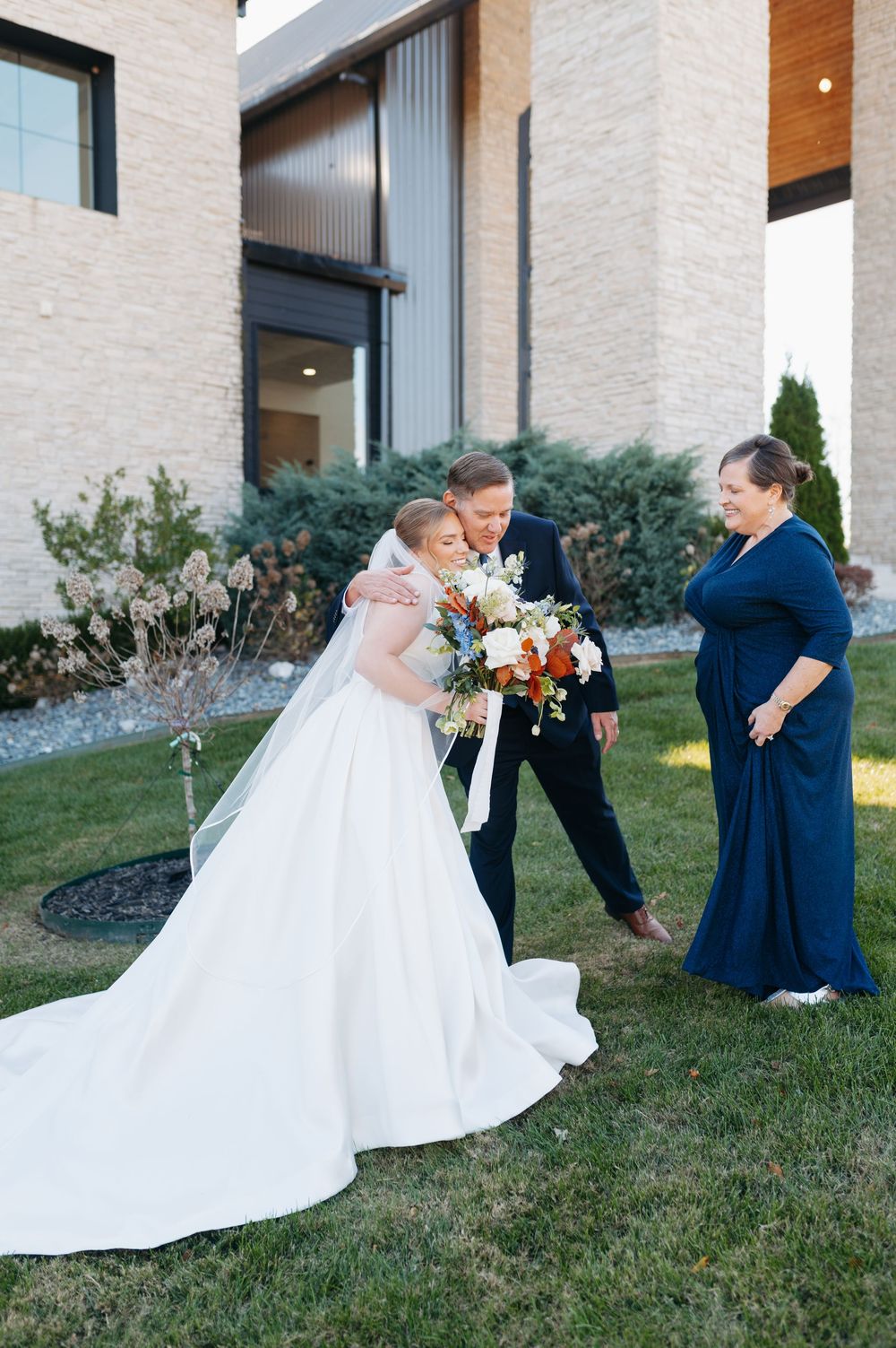 Bride and groom share a tender first look moment outdoors, as a woman in navy looks on, bride holding a colorful bouquet.