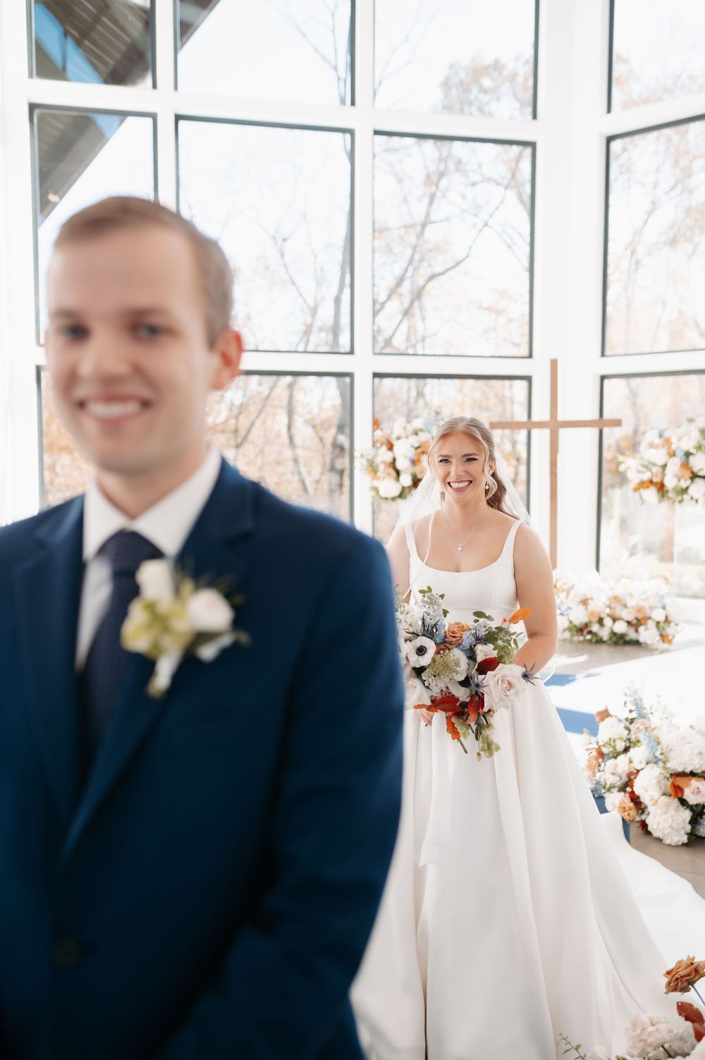 Bride in white gown holding floral bouquet smiles at groom in navy suit during wedding first look in chapel.