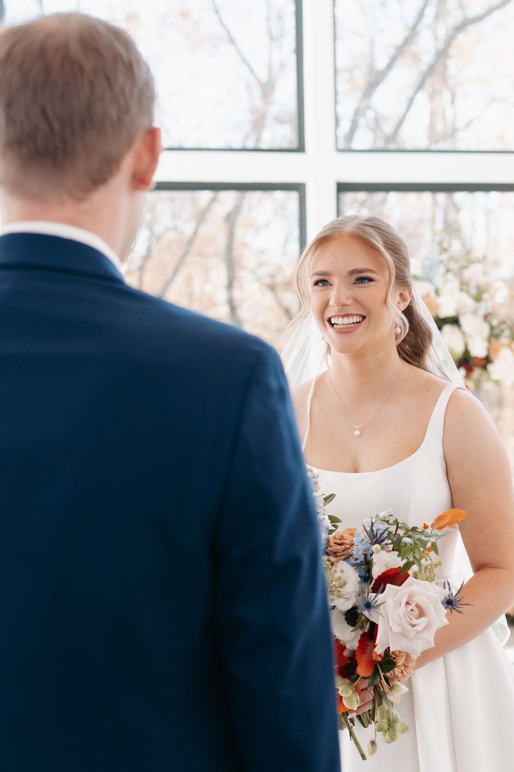 Smiling bride in white gown holds colorful bouquet facing groom in navy suit at bright windowed ceremony.