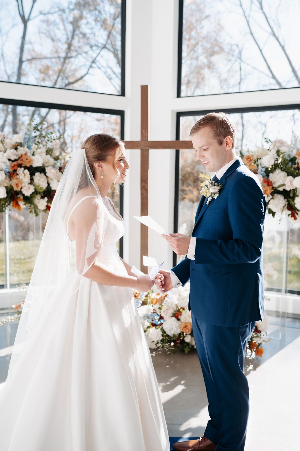Bride and groom exchange vows at altar with wooden cross and floral arrangements in sunlit wedding chapel.