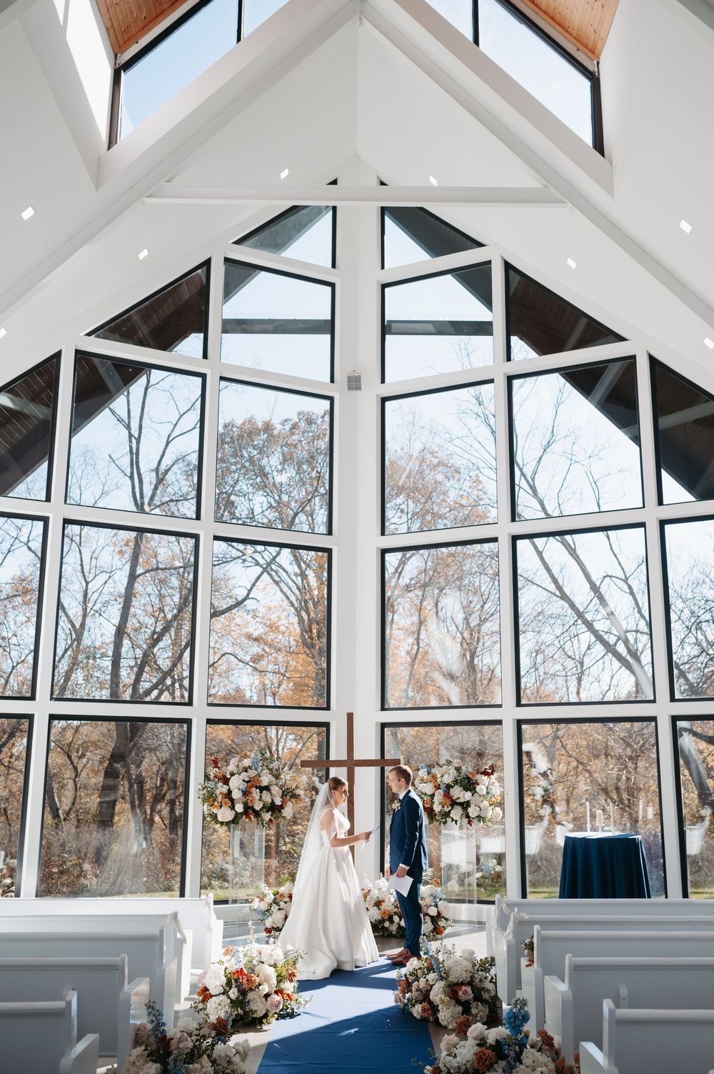 Bride and groom stand at the altar of a stunning glass chapel with soaring ceilings and floral arrangements.
