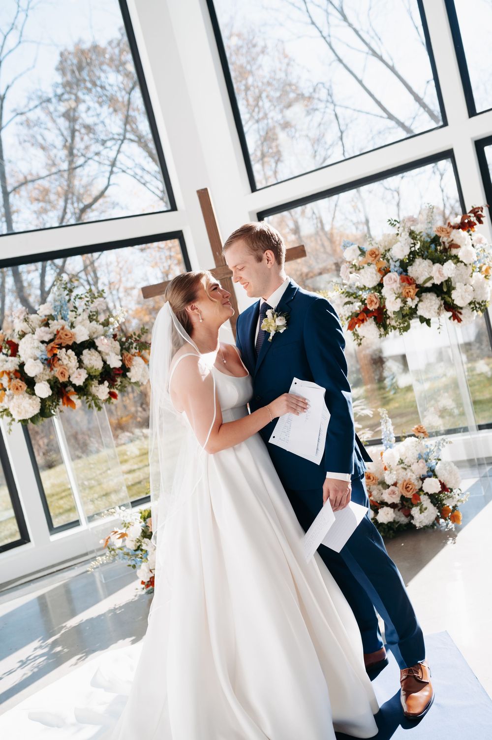 Bride and groom share an intimate moment at altar, surrounded by lush floral arrangements in a sunlit chapel.