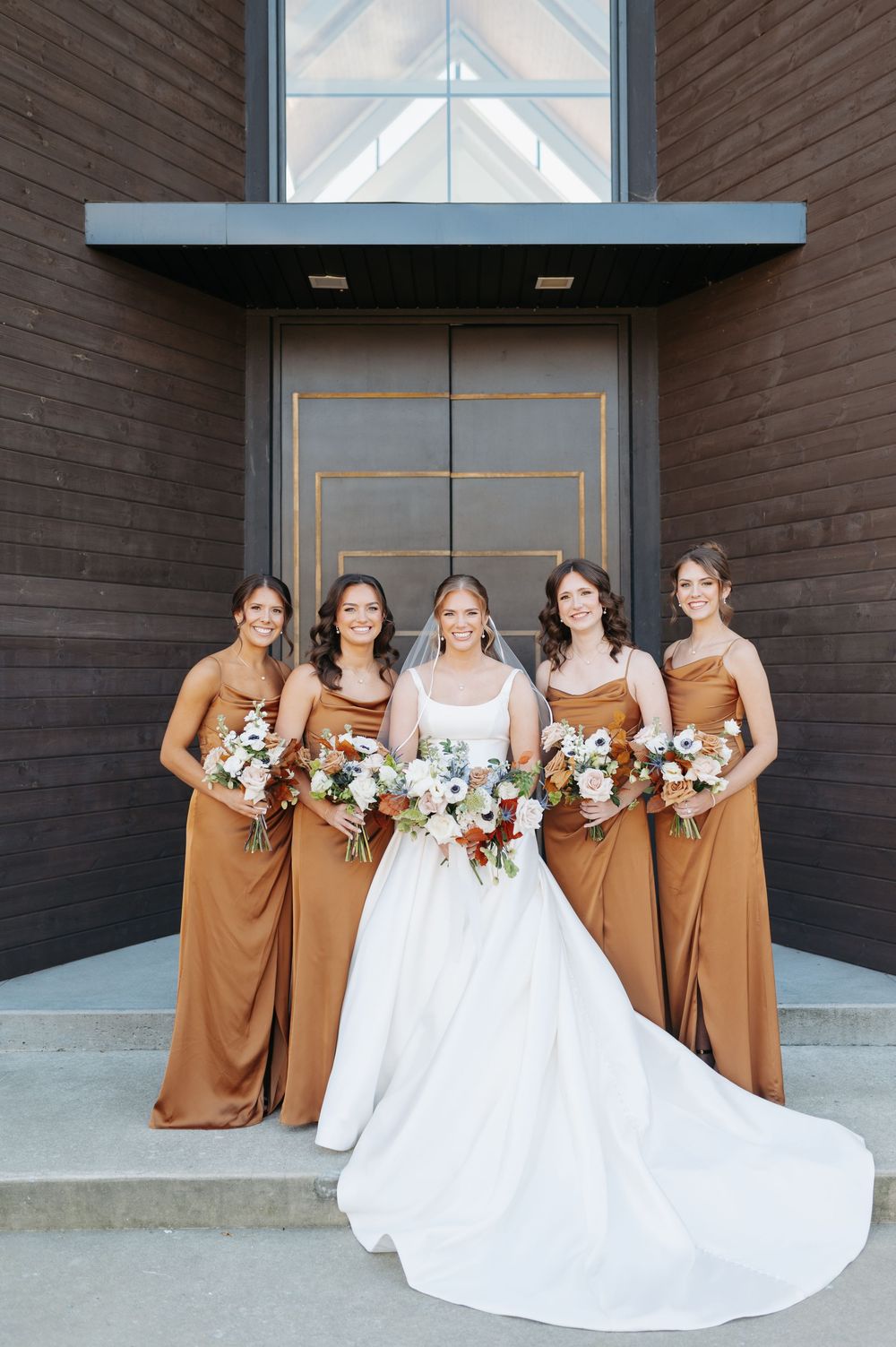 Bride in white gown poses with four bridesmaids in copper satin dresses holding bouquets.