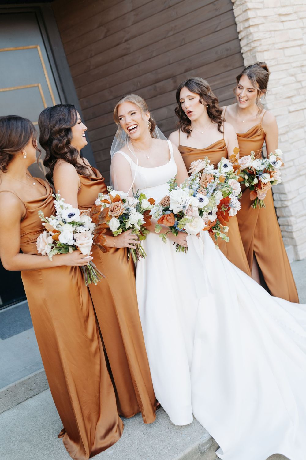 Bride in white gown laughs with bridesmaids in rust satin dresses holding floral bouquets.