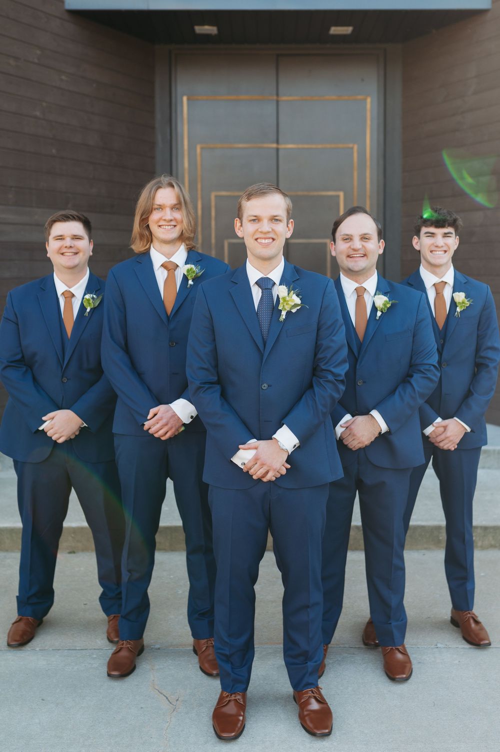 Five groomsmen in navy blue suits with brown ties and boutonnieres posing in front of a modern building.