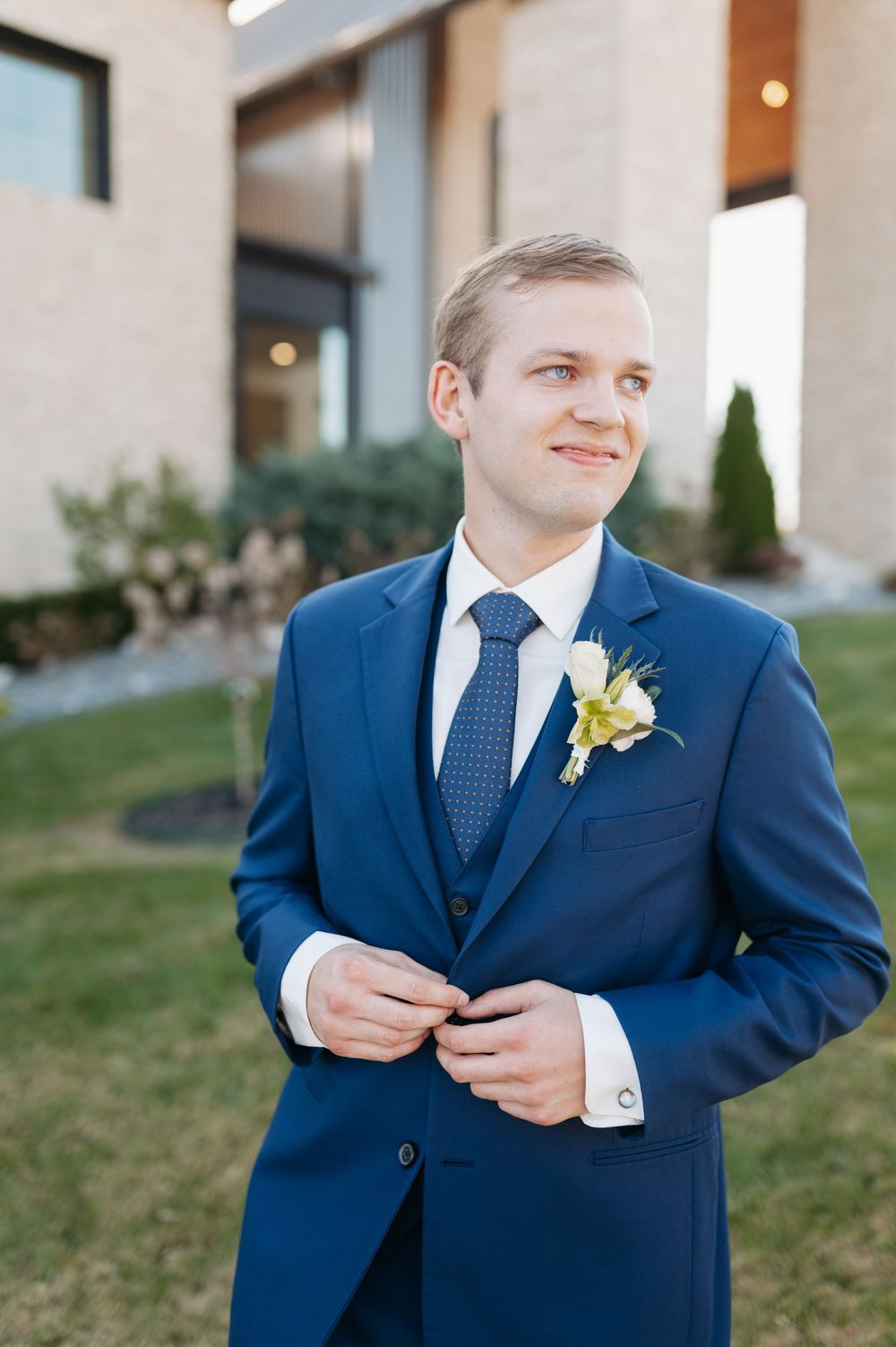 Dapper groom in navy blue suit adjusts jacket button outdoors before wedding ceremony.