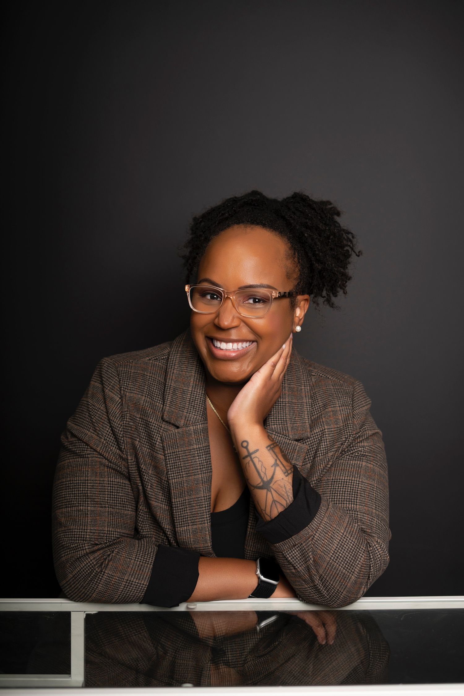 Smiling Black woman with natural hair, glasses, and plaid blazer leaning on white railing against dark background.