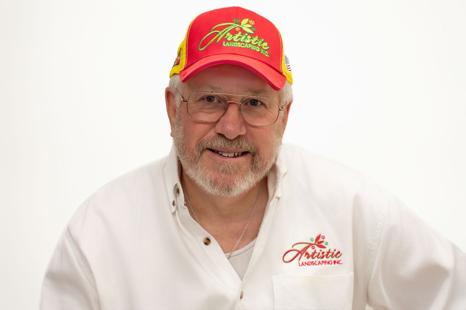 Older man smiling in a white Artistic Landscaping shirt and red branded cap.
