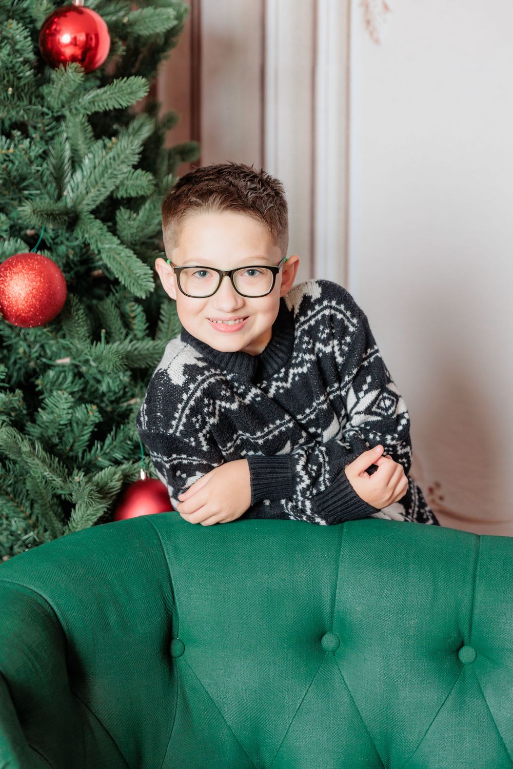 Smiling boy in Nordic sweater and glasses poses by decorated Christmas tree on green velvet chair.