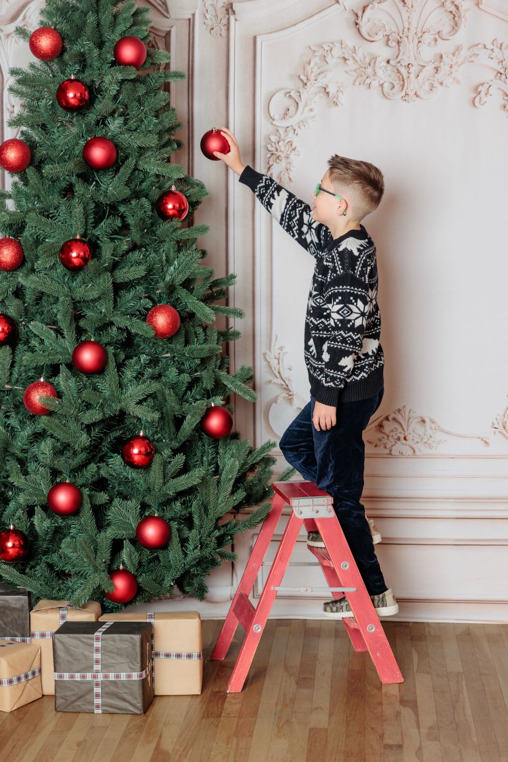Boy in Nordic sweater standing on red ladder decorating Christmas tree with red ornaments, gifts below.