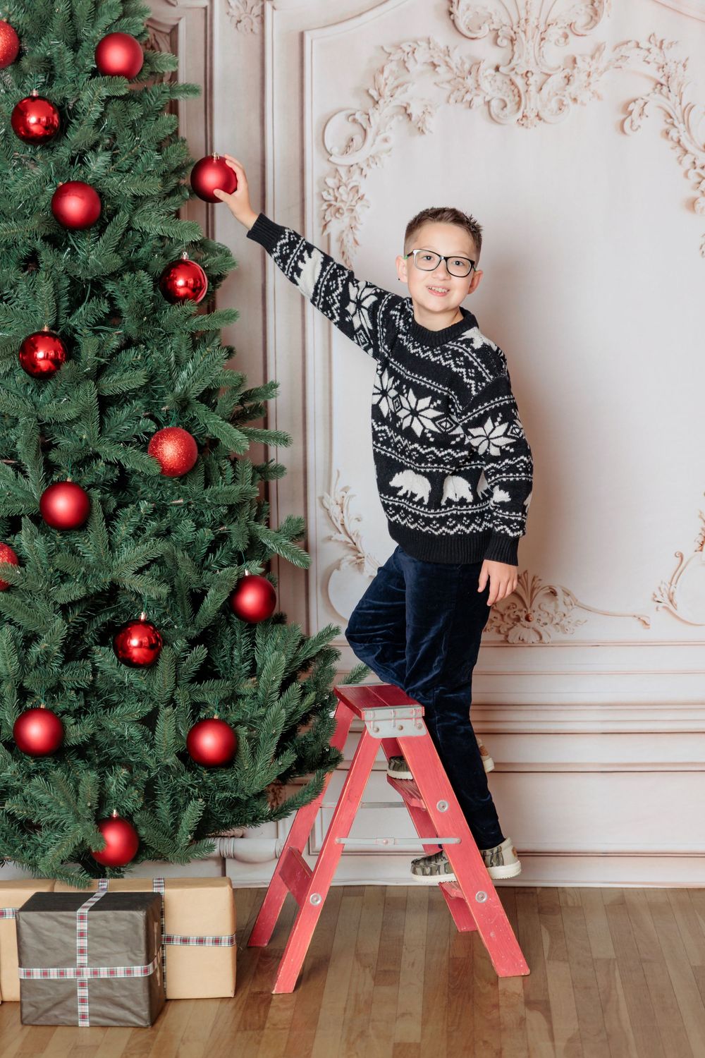 Smiling boy wearing glasses on red ladder placing red bauble on decorated Christmas tree with gifts.
