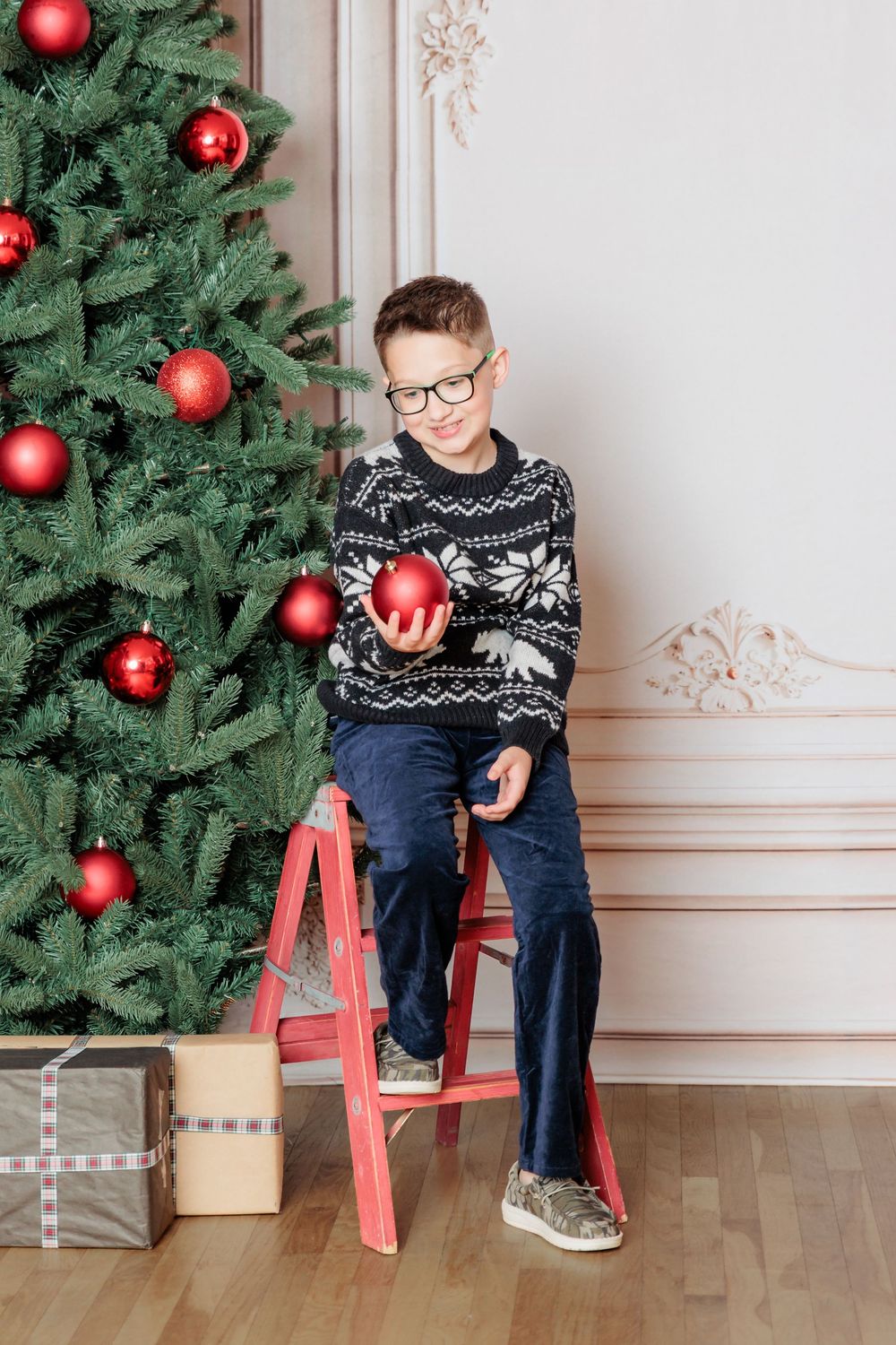 Boy in Nordic sweater sitting on red ladder holding Christmas ornament near decorated tree with gifts.