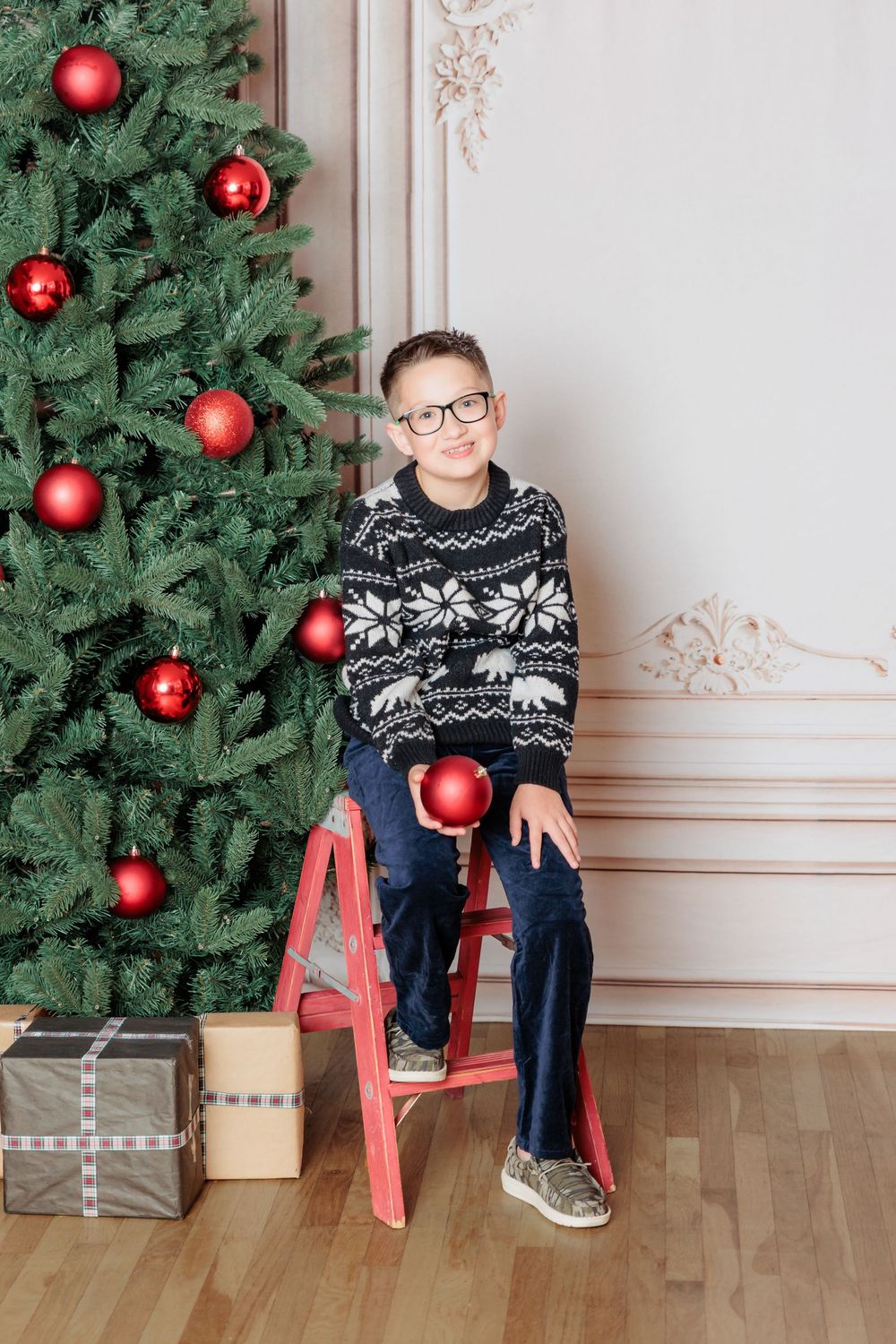 Boy in Nordic sweater and glasses sits on red ladder stool beside decorated Christmas tree holding red ornament.
