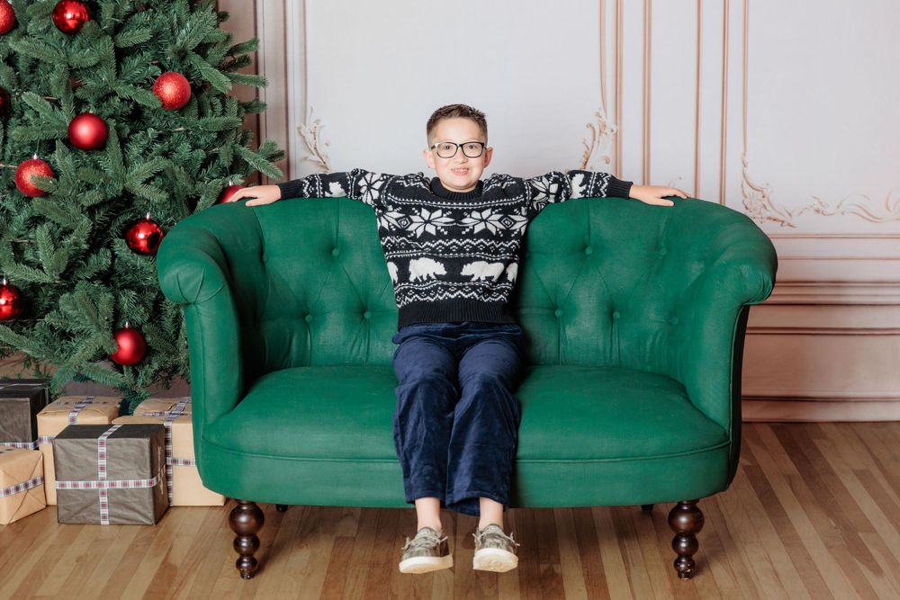 Boy in holiday sweater sitting on green tufted sofa beside decorated Christmas tree with red ornaments and gifts.
