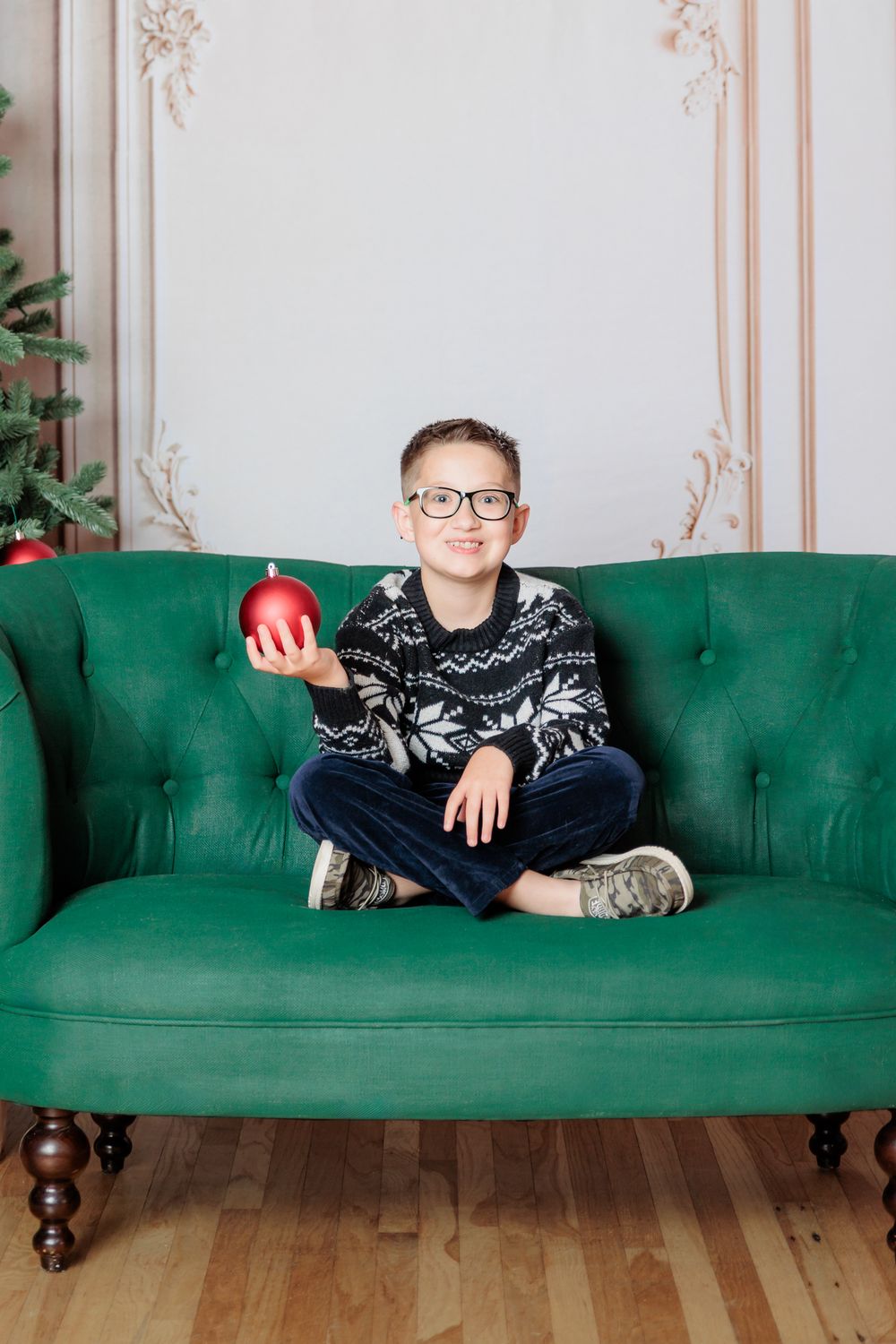 A smiling boy in a Nordic sweater sits cross-legged on a green sofa, holding a red Christmas ornament ball.