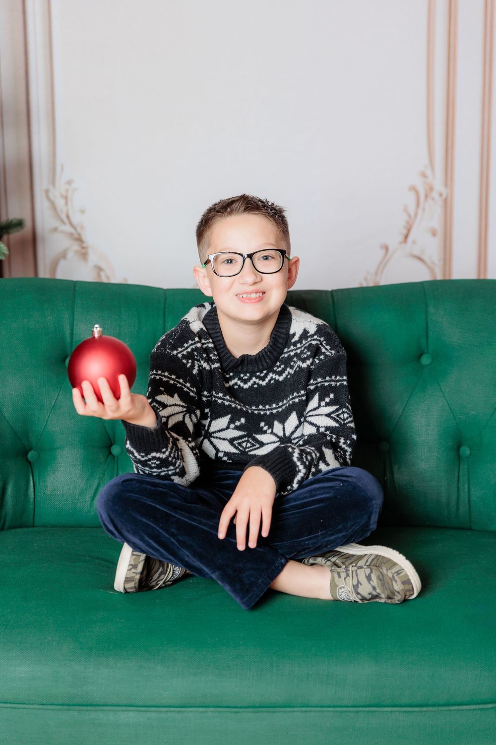 Smiling boy in glasses and Nordic sweater sitting cross-legged on green sofa holding red Christmas ornament ball.