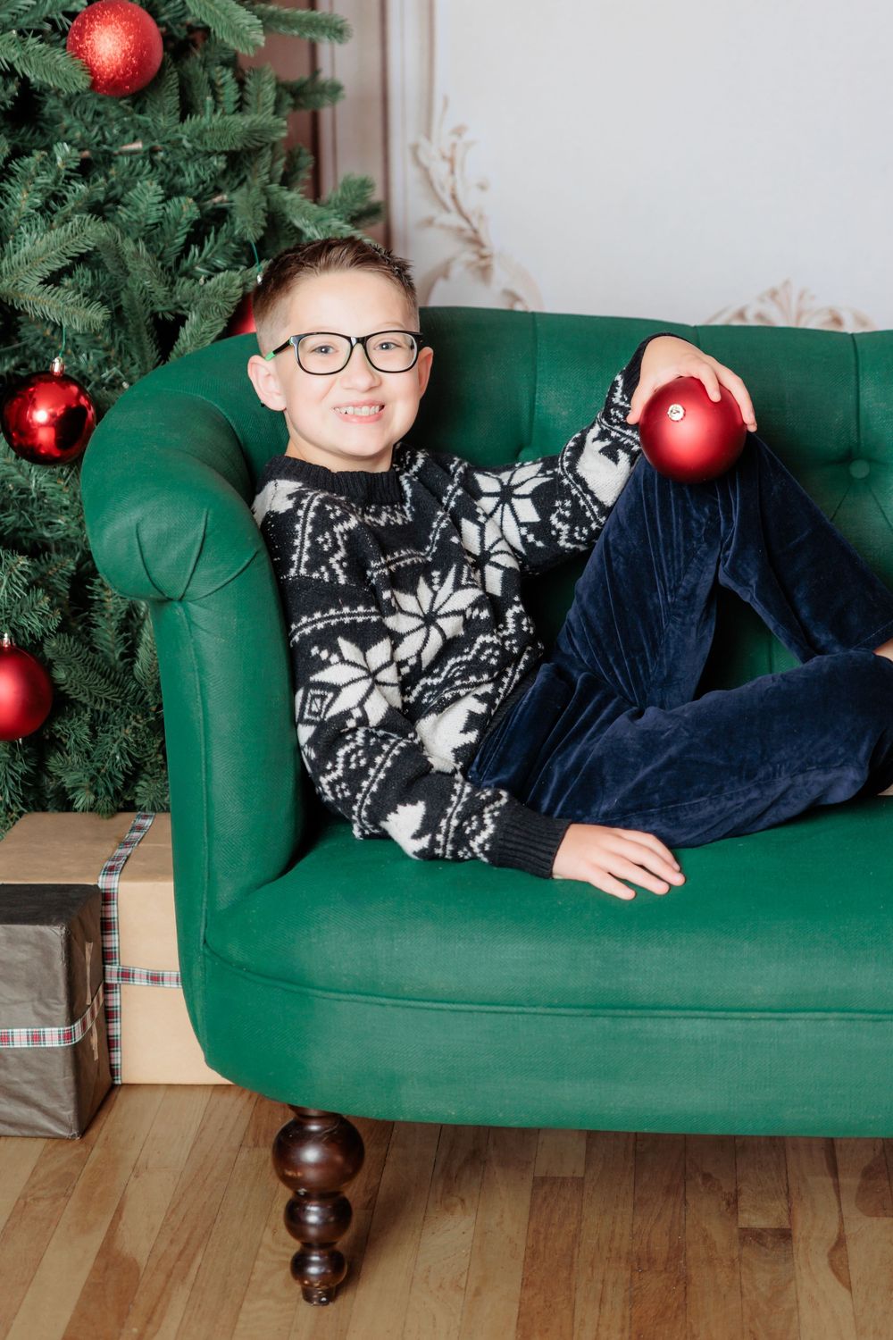 Smiling boy in Nordic sweater and glasses relaxing on green sofa holding red Christmas ornament near decorated tree.