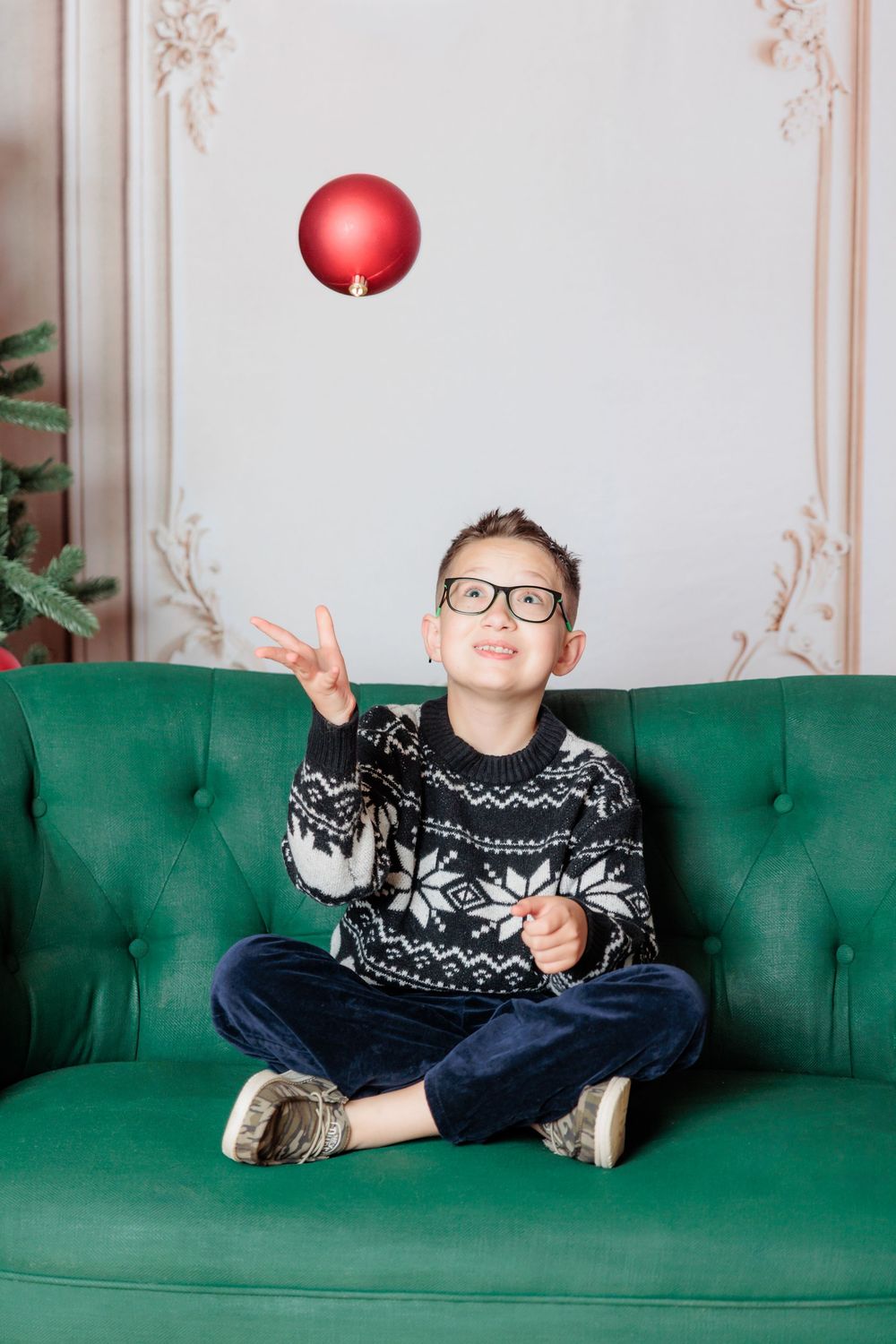 Boy in holiday sweater and glasses sitting on green sofa tossing red Christmas ornament ball in air.
