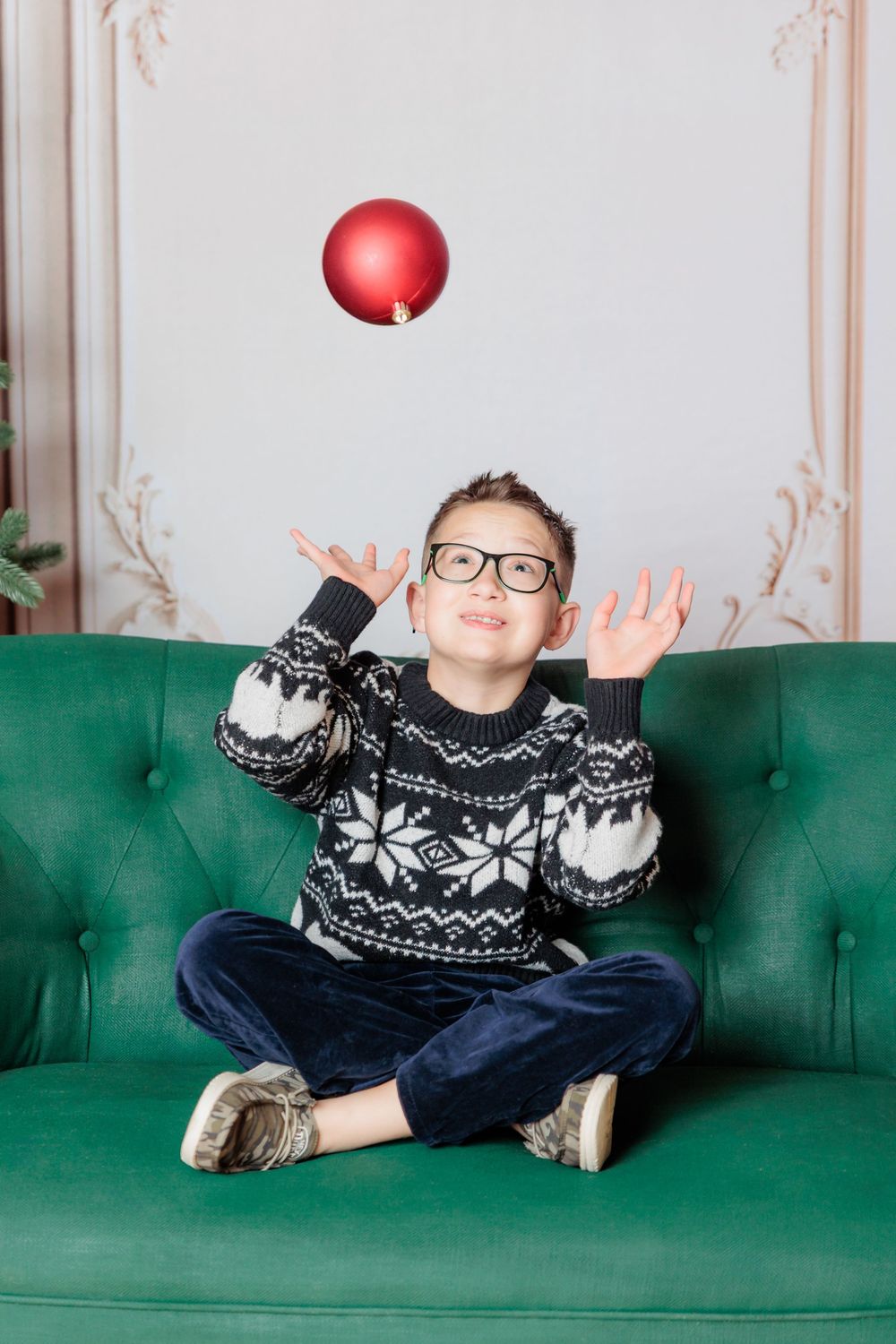 Boy in Nordic sweater and glasses sits cross-legged on green sofa, tossing a red Christmas ornament in the air.
