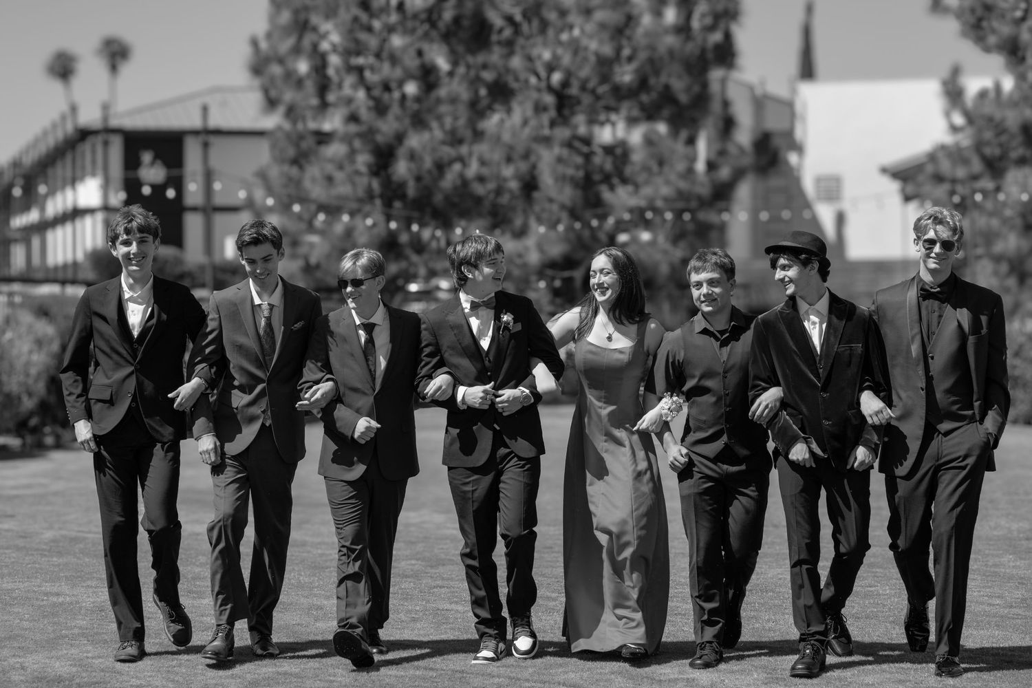 A group of seven teenagers dressed in formal attire walk together on a sunny lawn for prom. San Diego, CA
