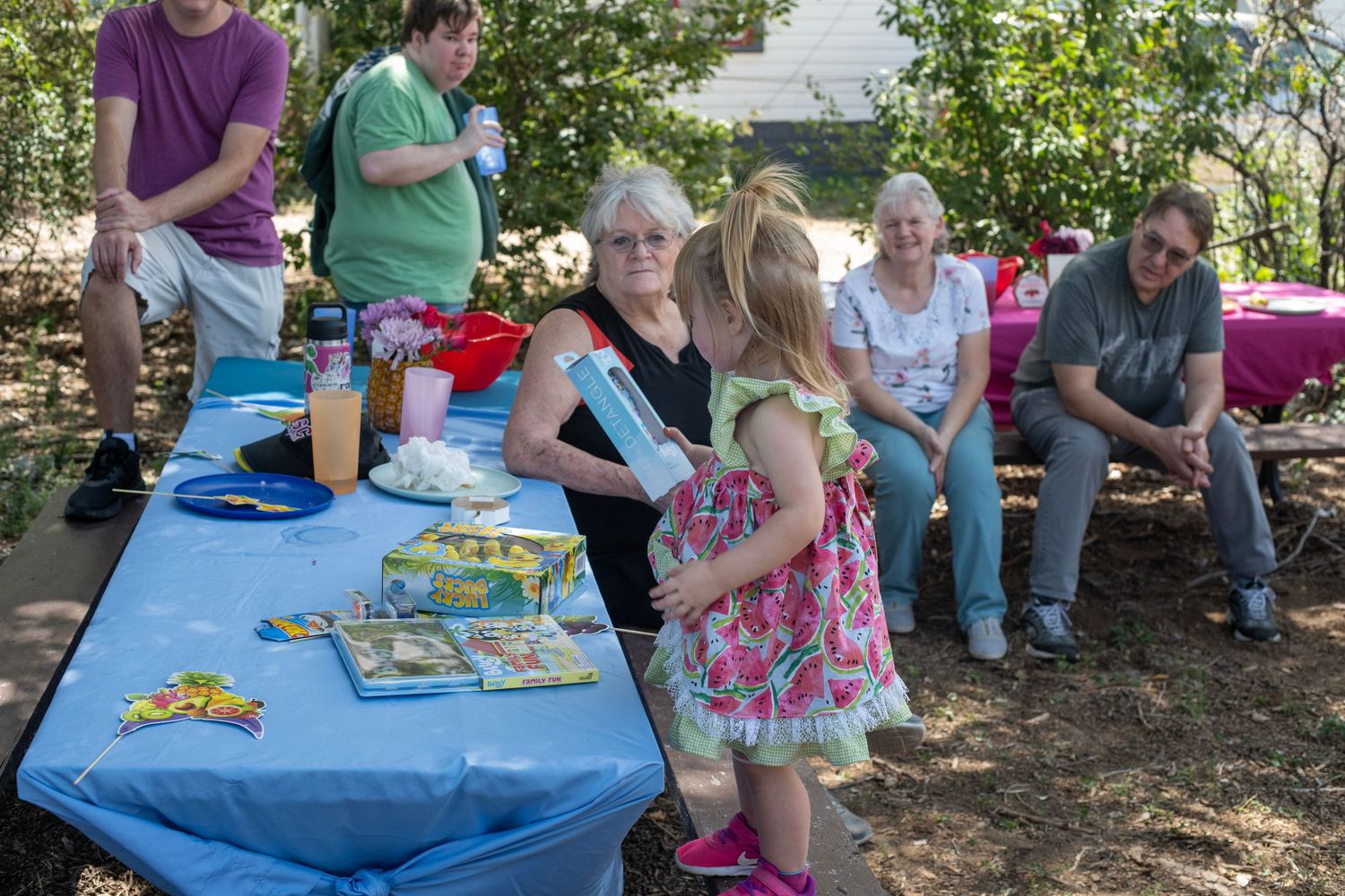 Outdoor birthday party with children and adults gathered around a blue table with gifts and decorations in a backyard.