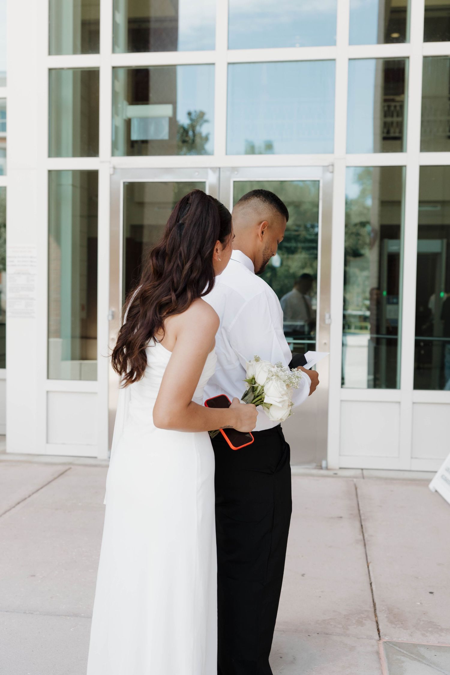 Bride in white dress holding bouquet embraces groom in white shirt and black pants outside glass building doors.