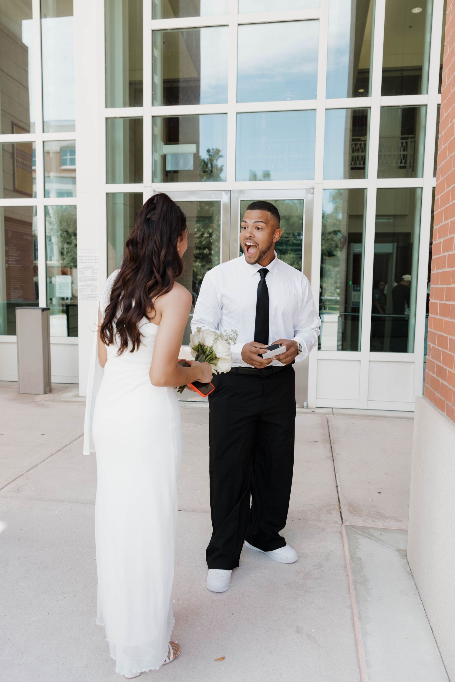 Groom reacts with surprise and joy seeing bride in white wedding dress during first look outside modern building.
