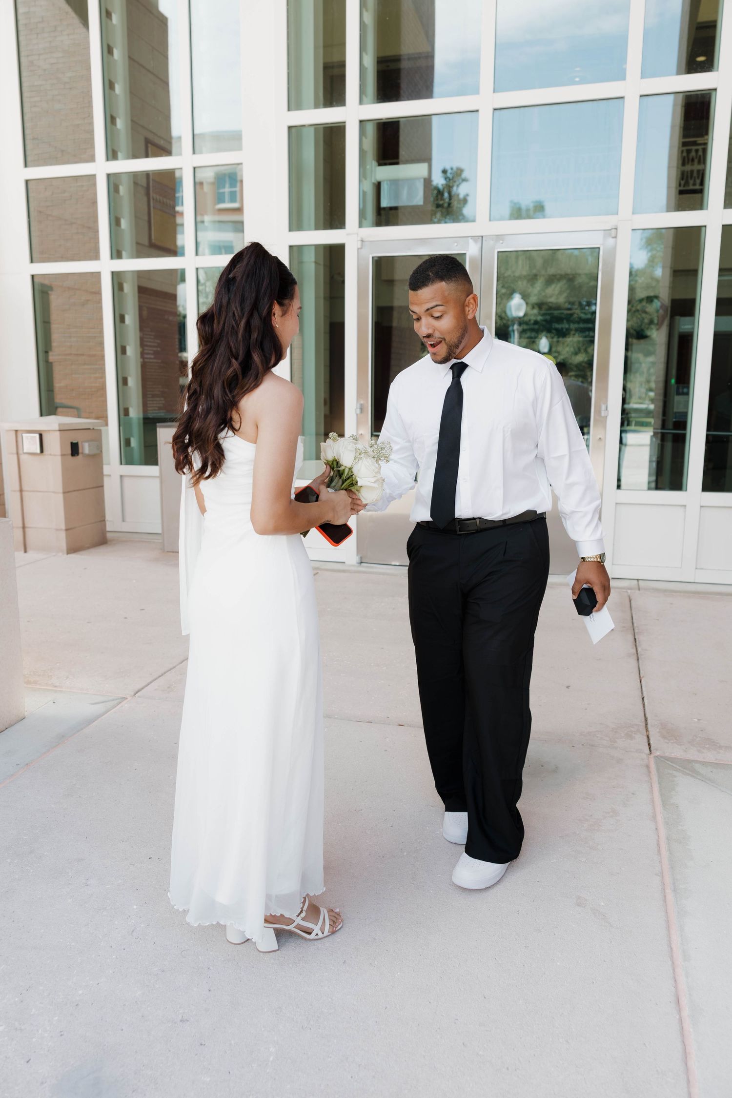 A bride in a white gown holds flowers while a groom in a white shirt and black tie smiles outside a building.