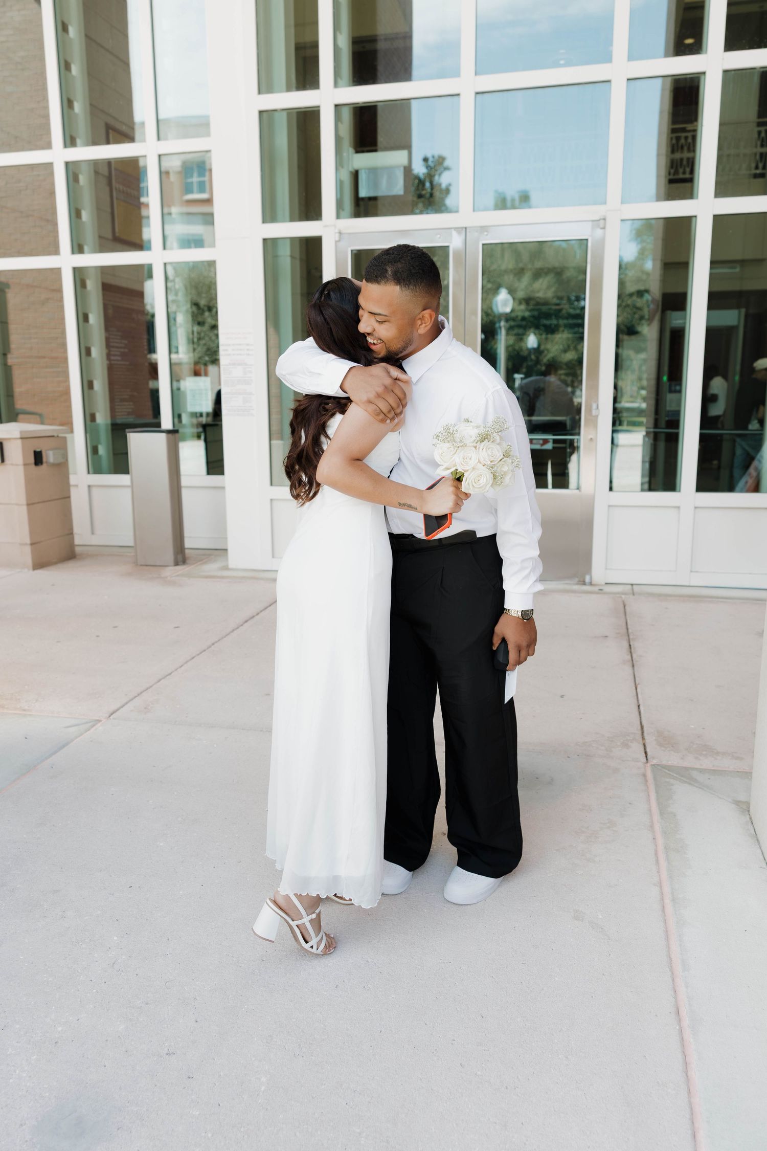 Bride in white dress and groom in black pants embrace outside a modern building, holding white flowers.