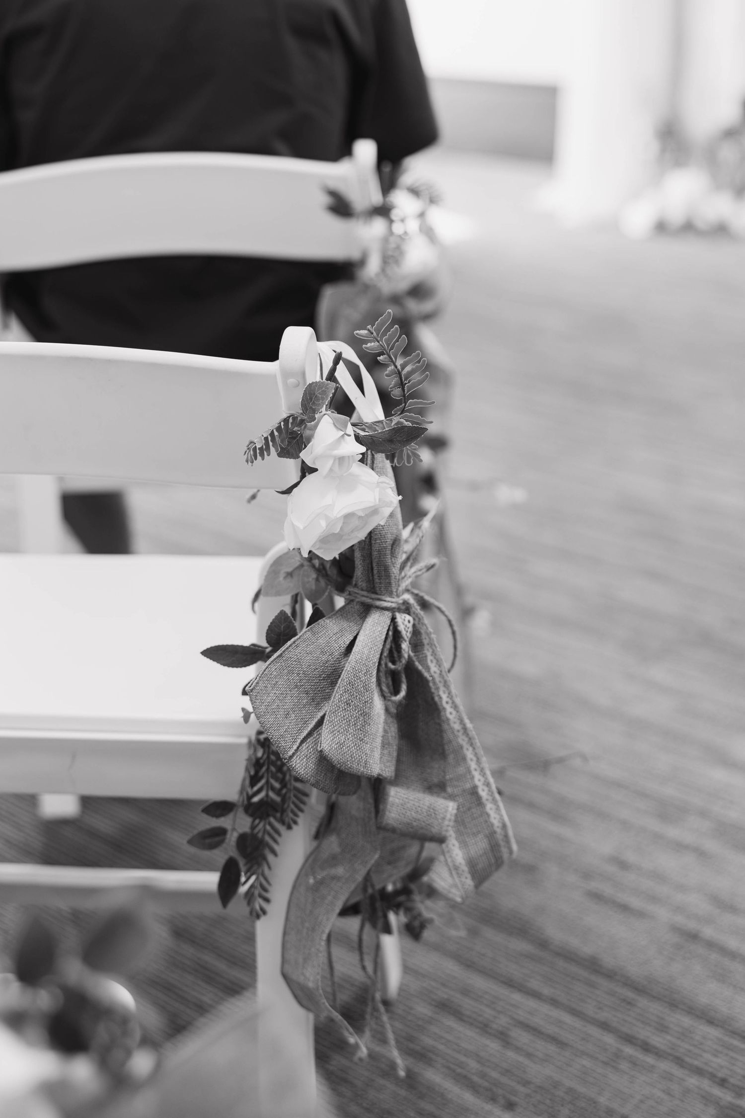 Black and white photo of white wedding chairs decorated with burlap bows and floral greenery arrangements along the aisle.