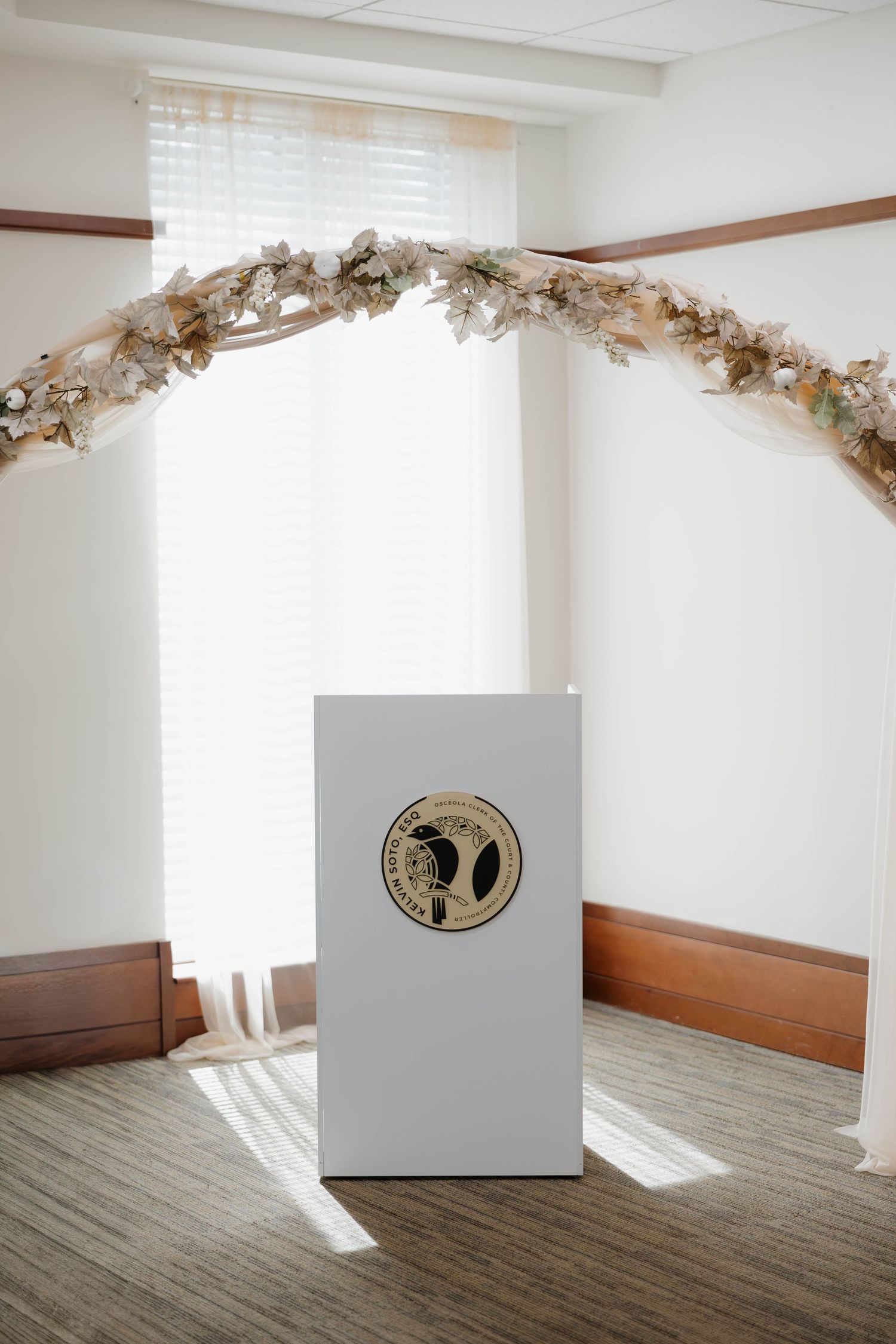 Elegant wedding ceremony arch with dried floral garland above white podium with gold seal in bright room.